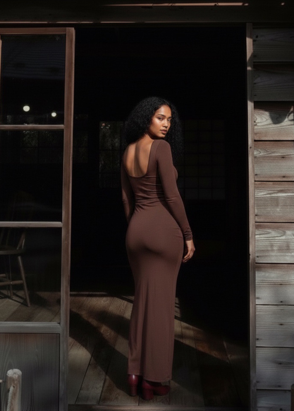 A woman stands in a doorway wearing a fitted, long-sleeve, backless maxi dress in a dark brown shade. The photo is taken at night with dramatic side lighting, casting a shadow on the rustic wooden wall and floor.
