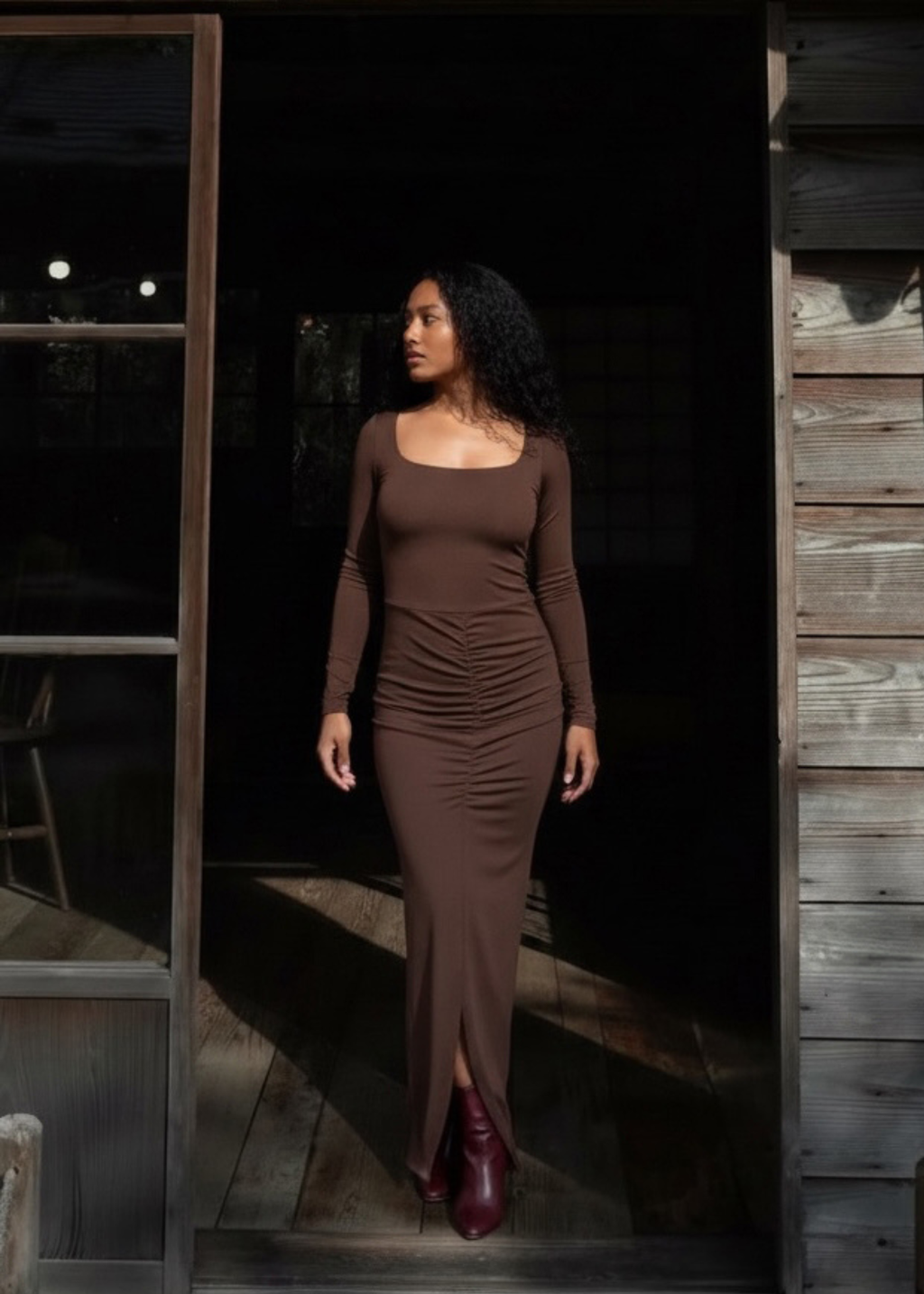A woman wearing a fitted long-sleeve brown maxi dress with ruching and a square neckline, standing in a doorway between dark wood and glass panels.